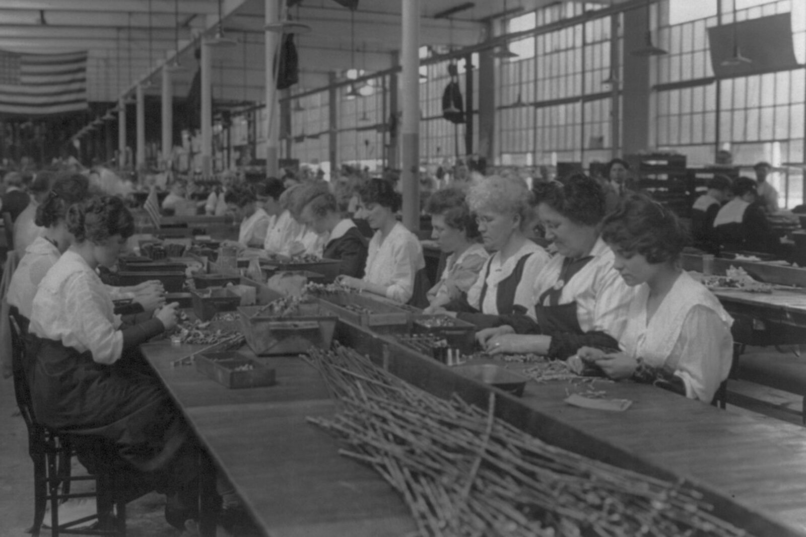 Black and white photo of women seated along a long factory table, inspecting small metal parts by hand, Hartford, 1914.
